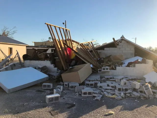 The image shows a pile of rubble in front of a house that has been torn down, with bricks, wooden...