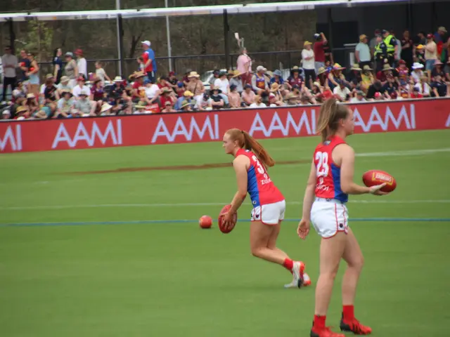 The image shows two women playing a game of AFL football on a field. They are both wearing sports...