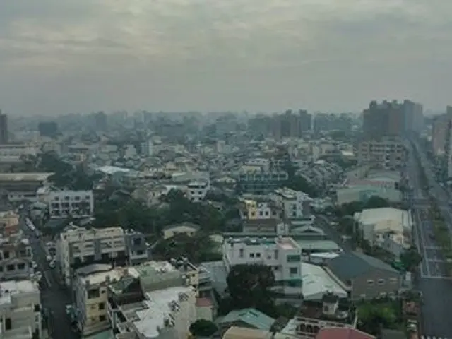 The image shows a view of a city from the top of a building, with many buildings, trees, and...