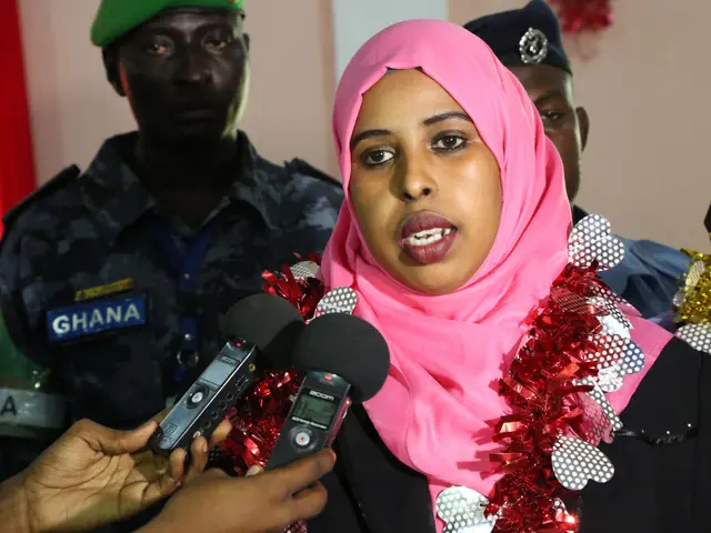 The image shows a woman in a pink headscarf speaking to reporters in front of a wall with a curtain...