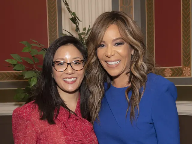 The image shows two women standing next to each other in front of a wall, both of them smiling. The...