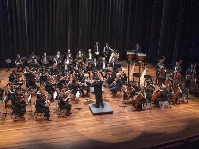 The image shows a large orchestra on a stage with a conductor in the center. The orchestra is...