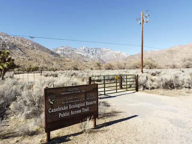 The image shows a sign on the side of a dirt road with mountains in the background. The sign reads...