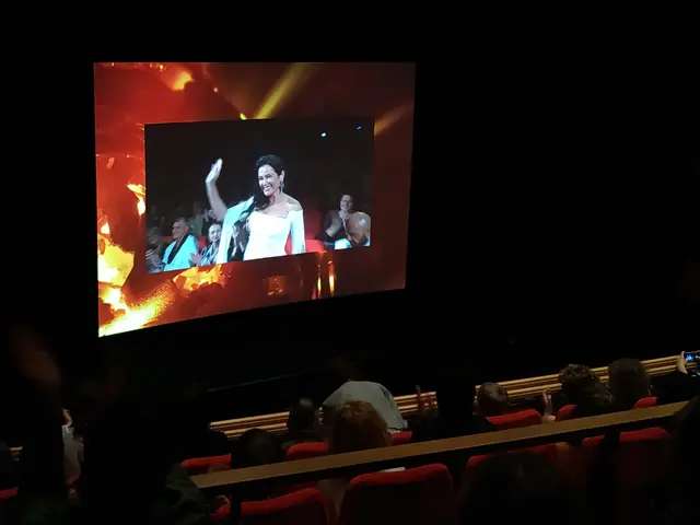 The image shows a group of people sitting in chairs in front of a large screen, with a dark...