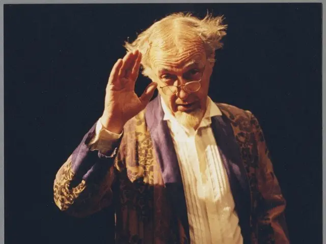 The image shows a man in a robe and glasses standing on a stage, with a dark background. He appears...