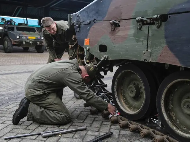 The image shows two men working on a military vehicle in a garage, surrounded by tools on the...
