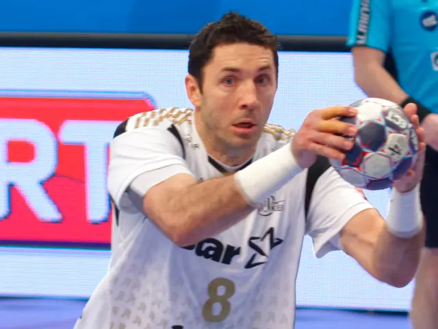 The image shows two men playing handball on a court, with one of them holding a ball in his hand....