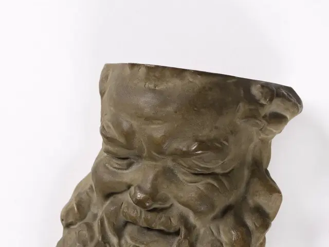 The image shows a bronze sculpture of a man's face with a beard, set against a white background....