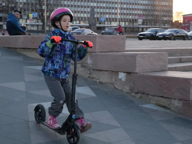 The image shows a young boy riding a scooter down a sidewalk, wearing a helmet and gloves. In the...
