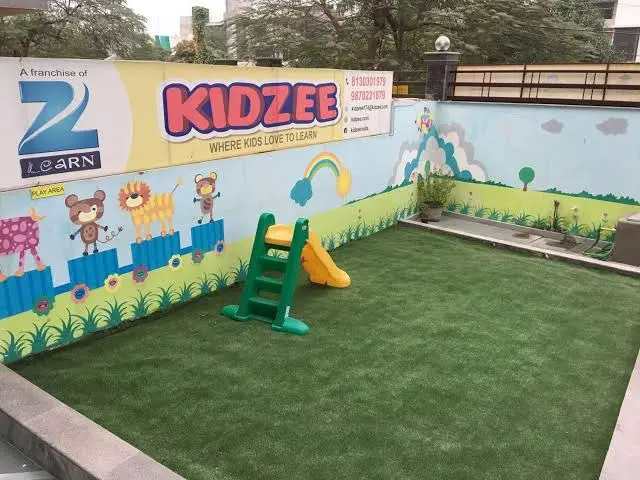 The image shows a kidzee play area with artificial grass, a slide, a wall with paintings and text,...