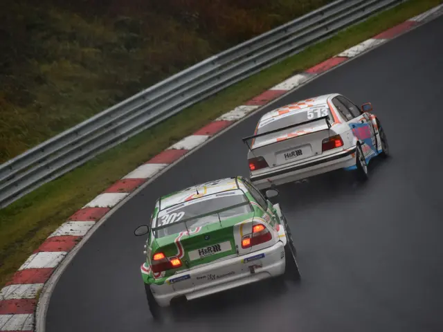 The image shows two cars racing on a race track in the rain, with a railing and grass on either...