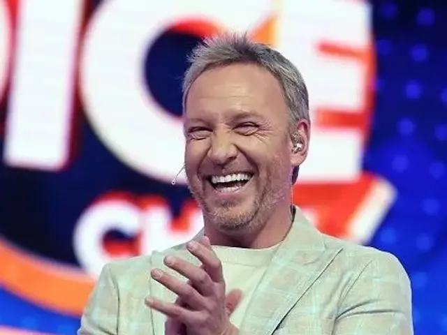 The image shows a man in a suit standing on a stage with a big smile on his face, clapping his...