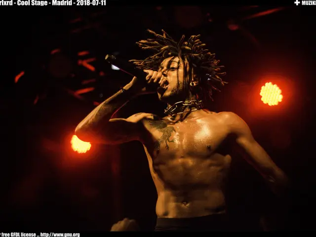 The image shows a shirtless man with dreadlocks singing into a microphone in front of a dark...