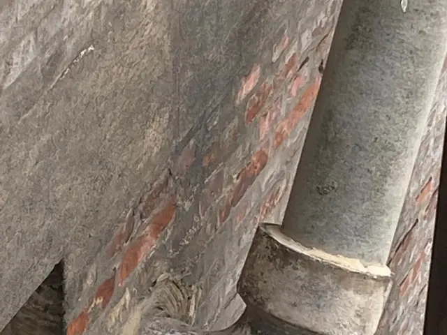 The image shows a close up of a metal pipe on the side of a building, with a wall in the...