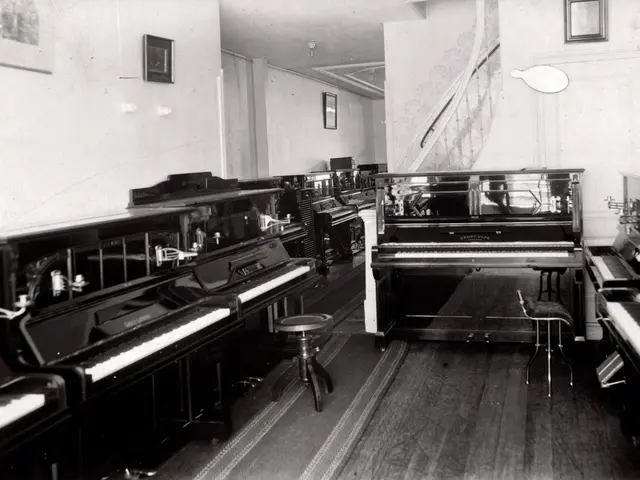 The image shows a black and white photo of a room filled with lots of pianos, stools, and frames...
