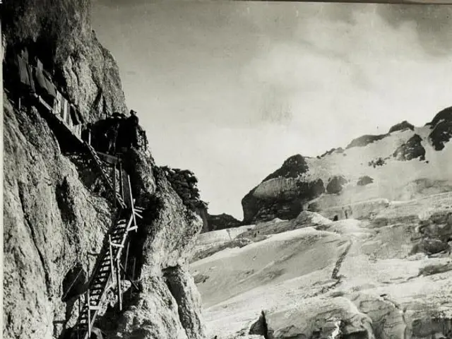 The image shows a black and white photo of a mountain with a few people climbing up the side of it,...