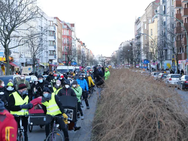 The image shows a large group of people wearing masks and safety vests riding bicycles down a...