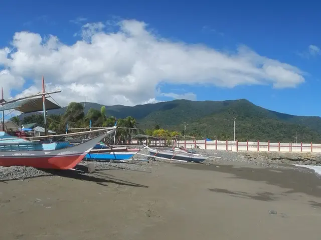 The image shows a beach with several boats sitting on top of it, surrounded by stones, poles, a...