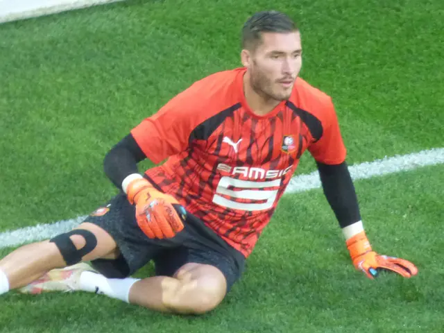 The image shows a soccer player sitting on the ground with his hands on his knees, wearing a red...