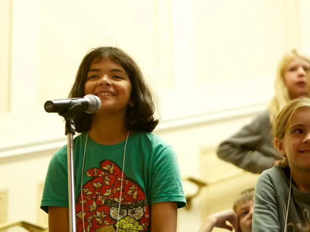 The image shows a young girl singing into a microphone in front of a group of children, with a wall...