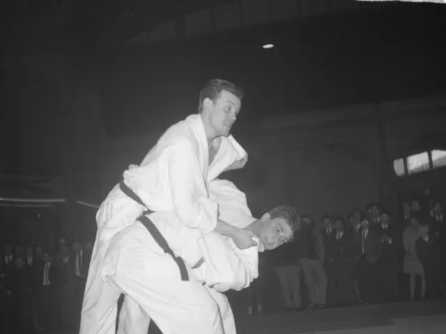The image shows two men in judo uniforms engaged in a fight in the center of the frame, with a...
