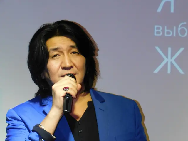 The image shows a man in a blue suit holding a microphone, standing in front of a screen with text...