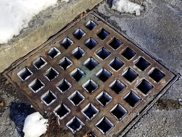The image shows a metal manhole cover on the side of a road, with snow on the ground around it.