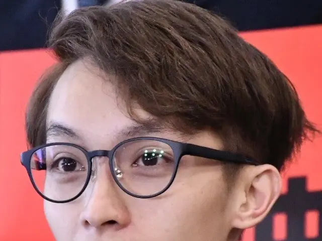 The image shows a man wearing glasses and a black shirt, holding a microphone, with a banner in the...