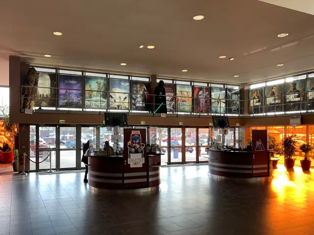 The image shows the lobby of a movie theater with a lot of people in it. There are houseplants,...