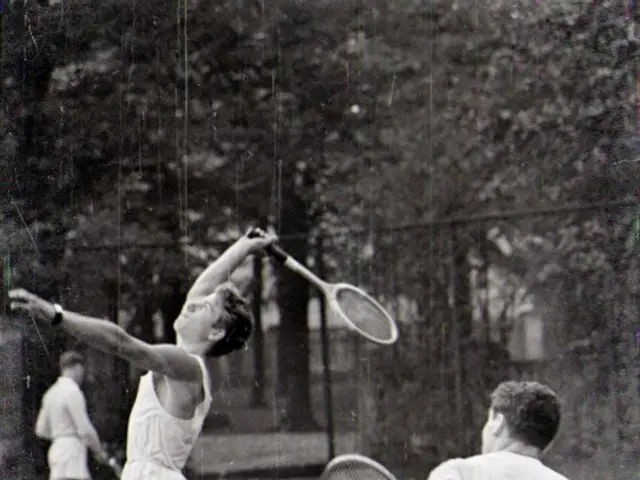 The image shows two men playing tennis on a court surrounded by trees. They are both holding...