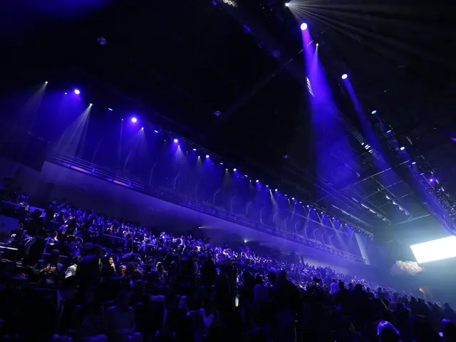 The image shows a large crowd of people sitting in front of a stage, illuminated by a few lights...