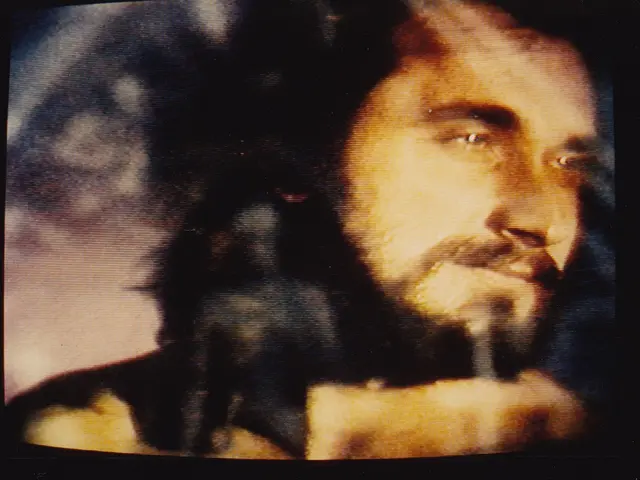 The image shows a man with a beard and mustache on a TV screen, with a dark background. His face is...