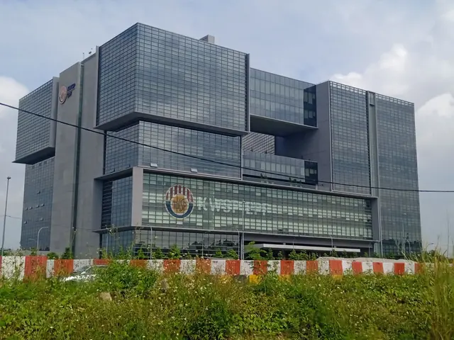 The image shows a large building with a lot of windows on the side of it, surrounded by plants, a...