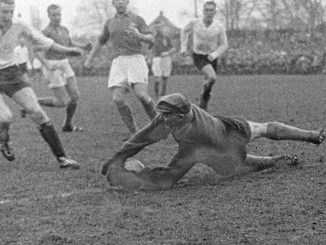 The image shows a group of men playing a game of rugby on a grassy field, with trees and a clear...