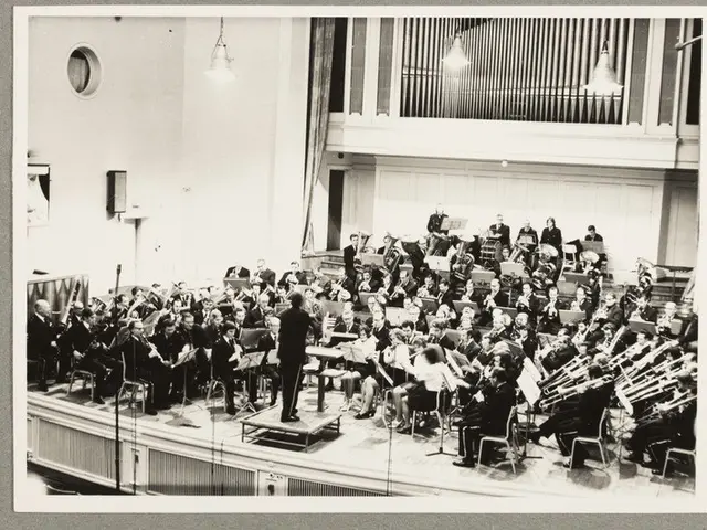 The image shows a black and white photo of an orchestra in a large room. There are many people...