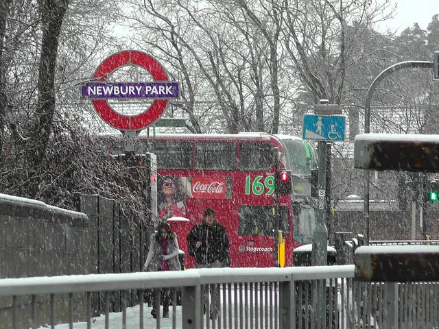 The image shows a red double decker bus driving down a snowy street next to a fence. There are...