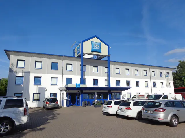 The image shows a building with windows and a signboard with text on it, a group of cars parked...