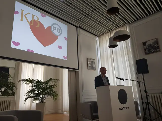 The image shows a man standing at a podium in front of a projection screen, with a microphone on...