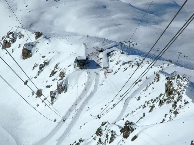 The image shows an aerial view of a ski lift going down a snowy mountain, with ropeway wires,...