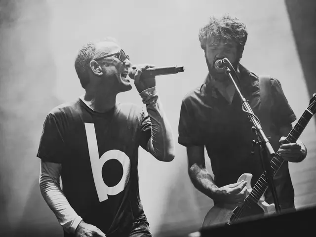 The image shows two men singing and playing guitars on stage at a concert. The man on the left is...
