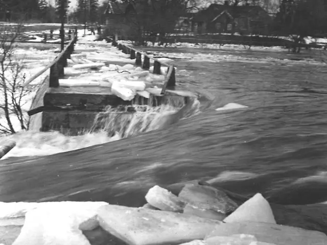 The image shows a black and white photo of a river with ice on it, surrounded by trees and houses...