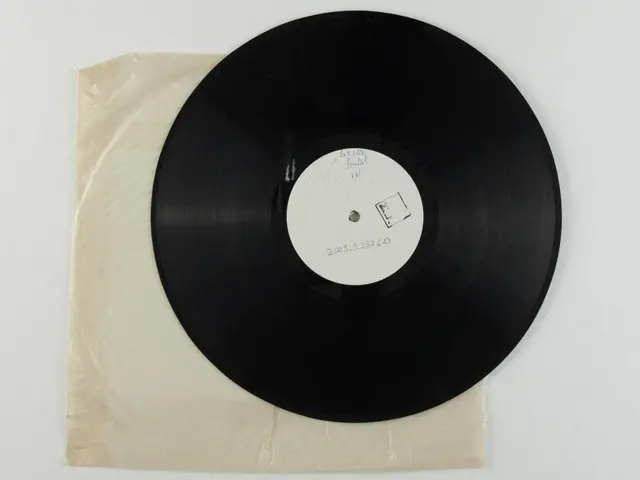 The image shows a black vinyl record sitting on top of a piece of paper on a white surface. The...