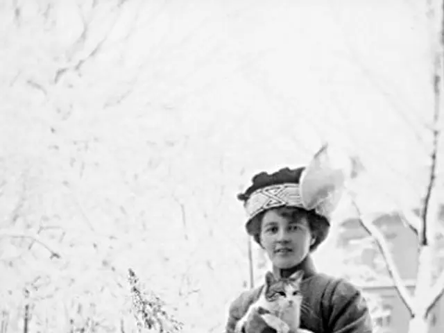 The image shows an old black and white photo of a woman standing in the snow, holding a cat in her...