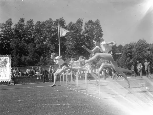 The image shows a black and white photo of a group of people jumping over hurdles on a track, with...