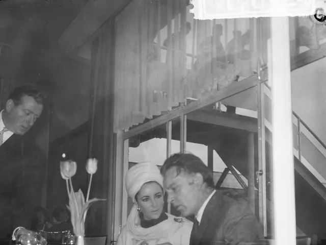 The image shows a black and white photo of a man and woman sitting at a table in a restaurant. On...