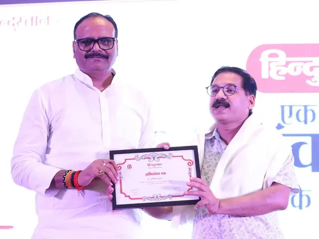 The image shows two men standing next to each other, both wearing spectacles and holding a...