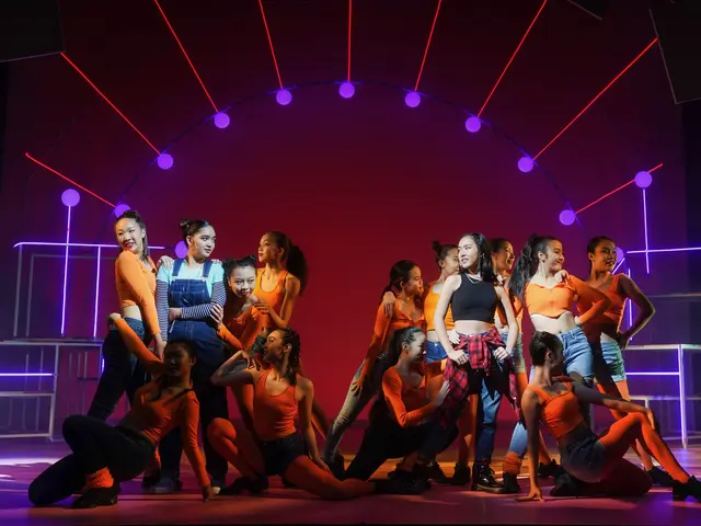 The image shows a group of young dancers on a stage, illuminated by colorful lights and surrounded...