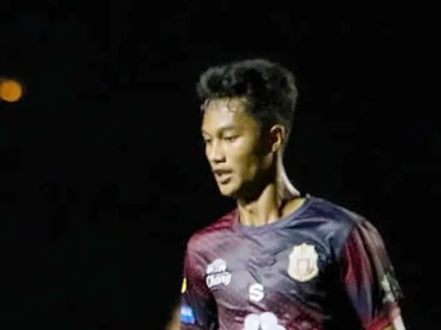 The image shows a man in a maroon shirt and red shorts playing soccer on a field, with another...