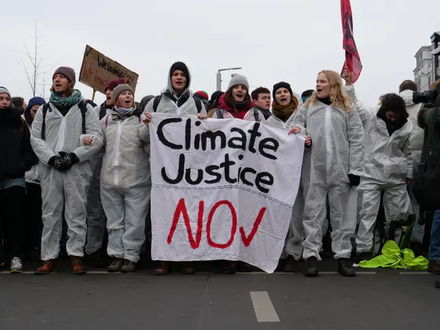 The image shows a group of people standing on the side of a road, holding a banner that reads...
