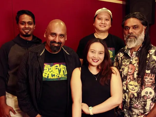 The image shows a group of people standing next to each other in front of a red wall, smiling. In...